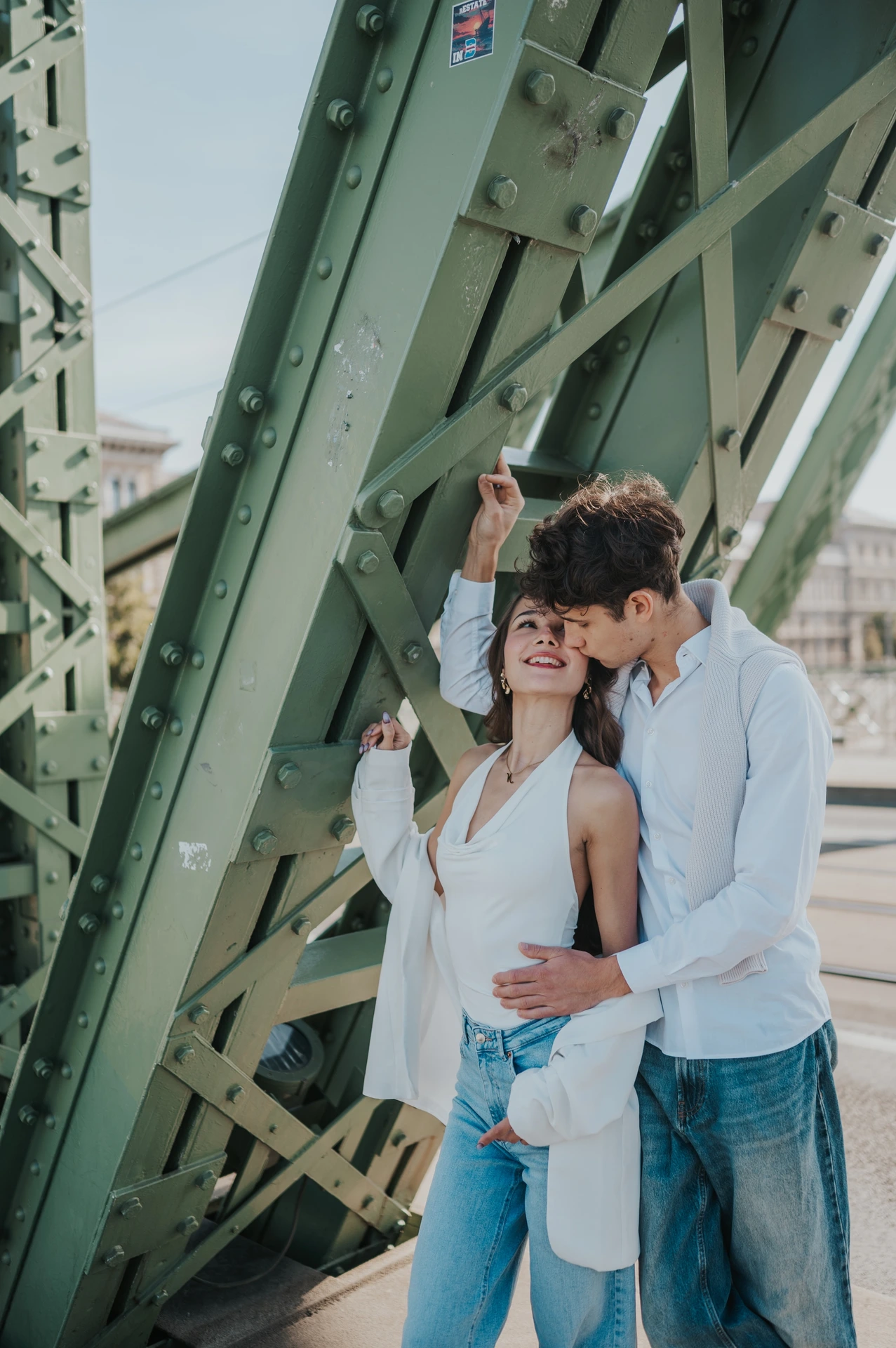 The couple smiling while standing close together beside the riveted green beams during their Budapest photoshoot on Liberty Bridge.