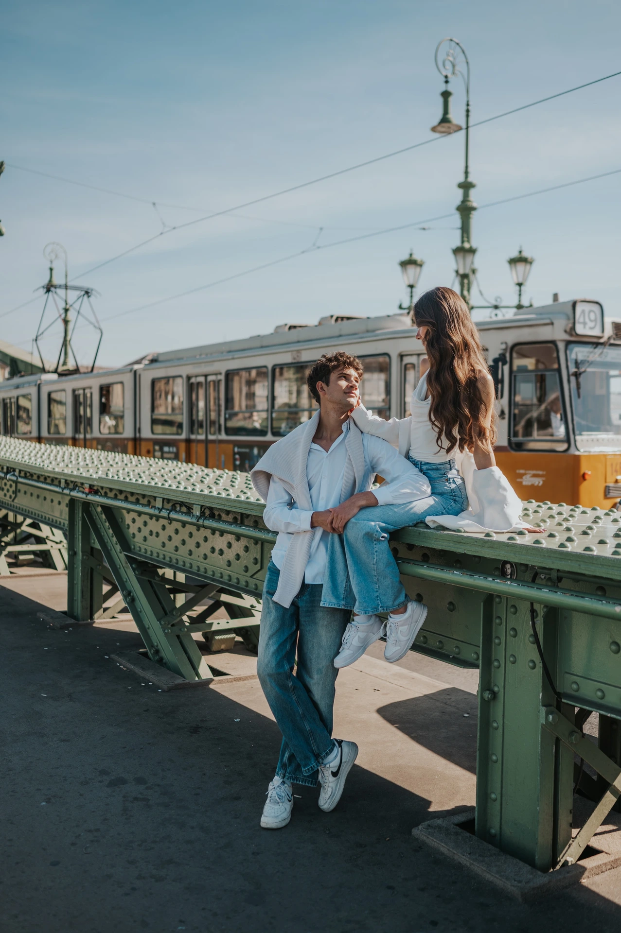 The couple standing close beside Liberty Bridge’s green railing during their Budapest photoshoot, with a decorative streetlamp and yellow tram behind them.