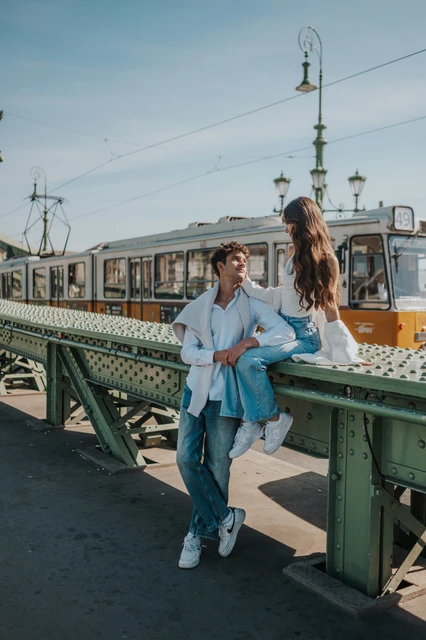 Budapest photoshoot with the Liberty Bridge streetlamp and a yellow tram The couple standing close beside Liberty Bridge’s green railing during their Budapest photoshoot, with a decorative streetlamp and yellow tram behind them.