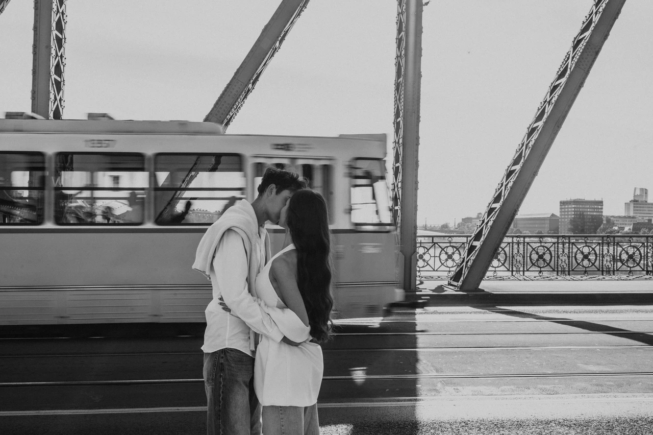 Budapest photoshoot with a Liberty Bridge tram in black and white Black-and-white photograph of the couple kissing during their Budapest photoshoot on Liberty Bridge, with a passing tram behind them.