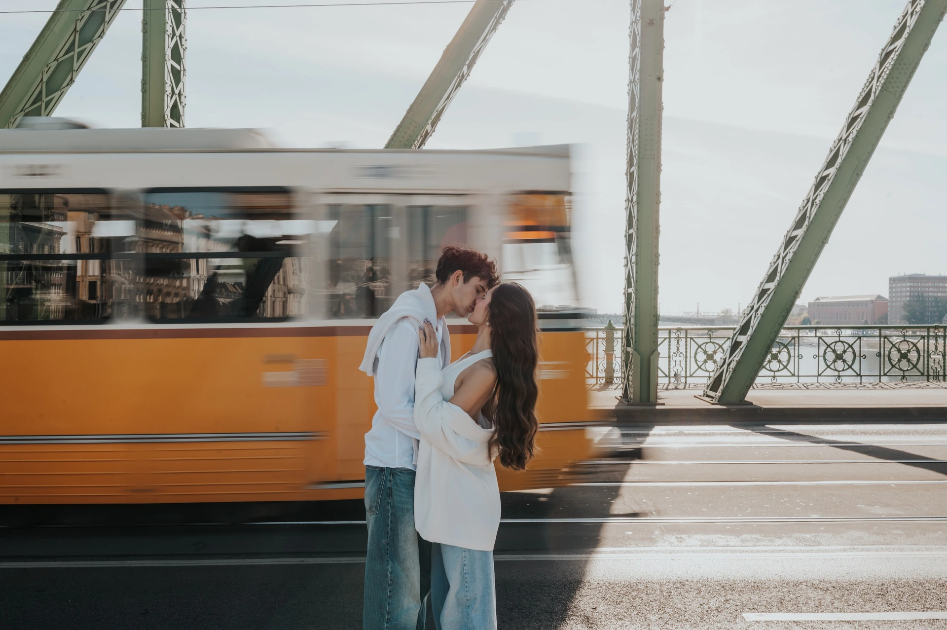 The couple holding each other close for a kiss during their Budapest photoshoot on Liberty Bridge, with the yellow tram blurred behind them.