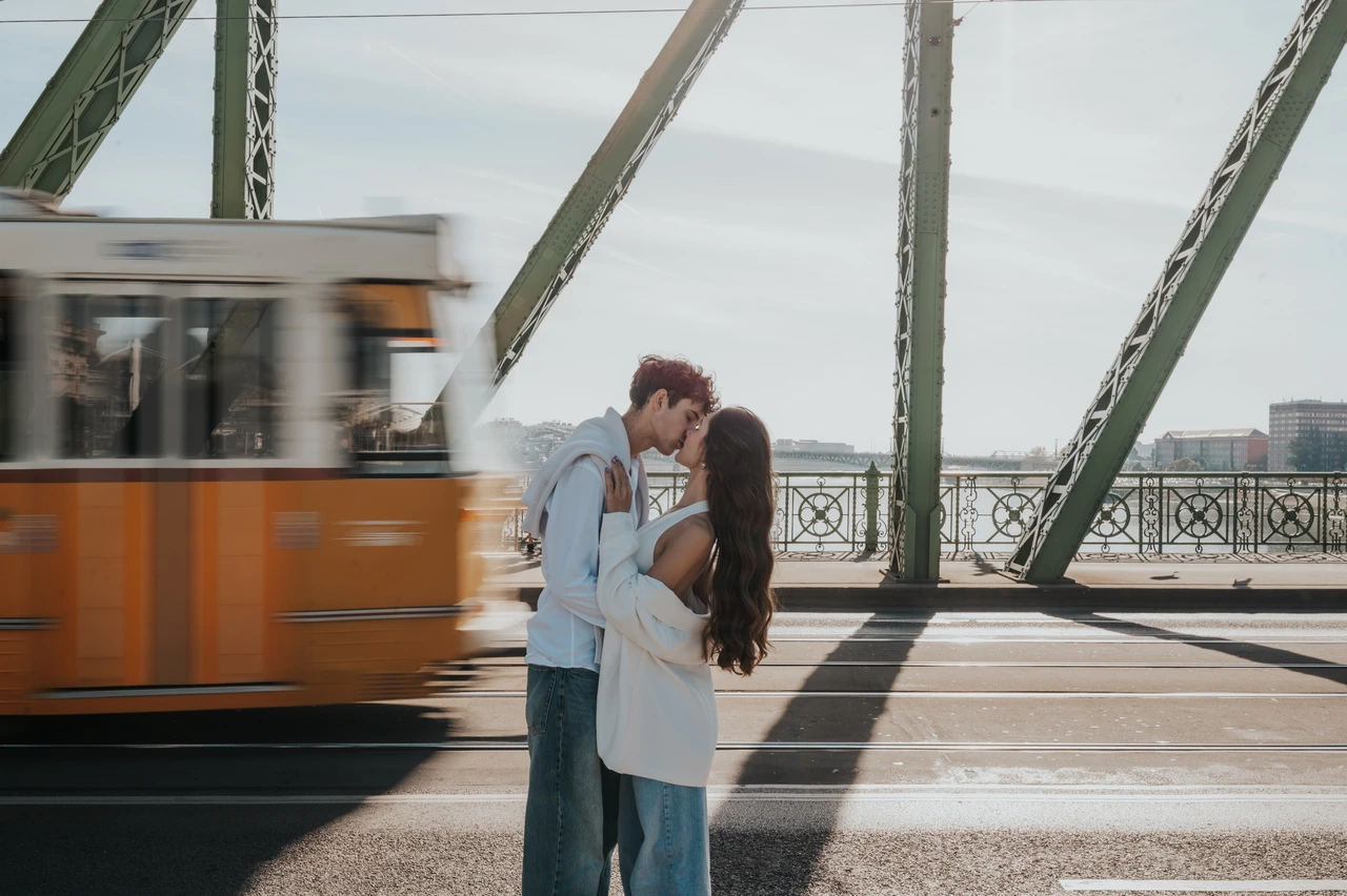Budapest photoshoot with a tram moving through Liberty Bridge Wide view of the couple embracing for a kiss during their Budapest photoshoot, with a yellow tram passing between Liberty Bridge’s green beams.