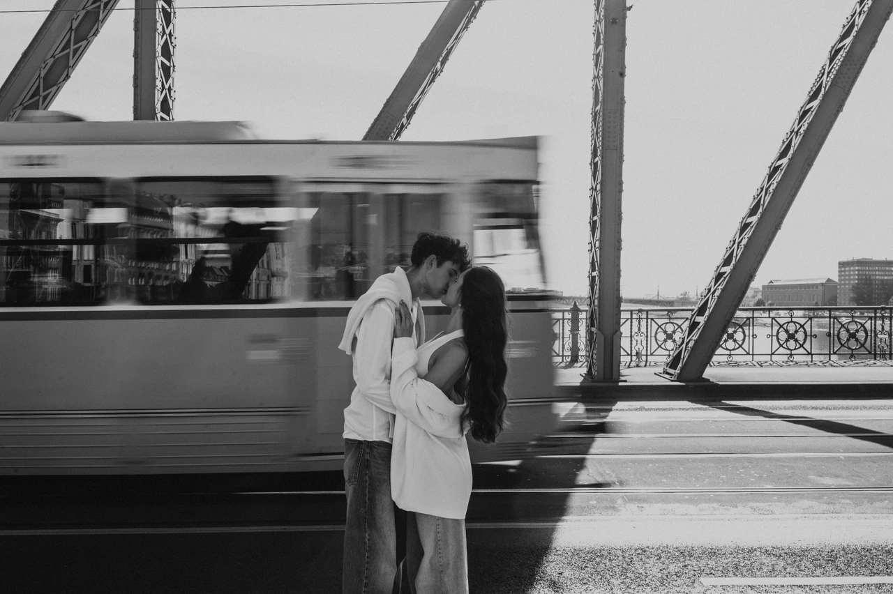 Couple photoshoot in Budapest beside a passing tram in black and white Black-and-white portrait of the couple kissing during their Budapest photoshoot on Liberty Bridge, with the motion of a passing tram in the backdrop.