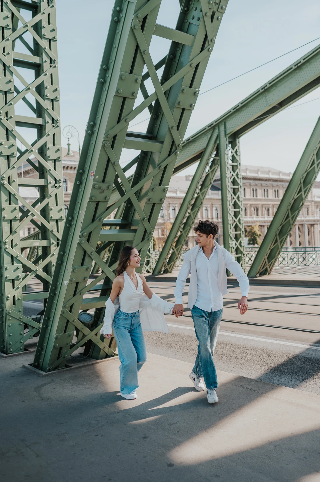 The couple walking hand in hand along Liberty Bridge during their Budapest photoshoot, framed by its tall green steel structure.