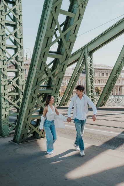 Budapest couple photoshoot walking along Liberty Bridge The couple walking hand in hand along Liberty Bridge during their Budapest photoshoot, framed by its tall green steel structure.