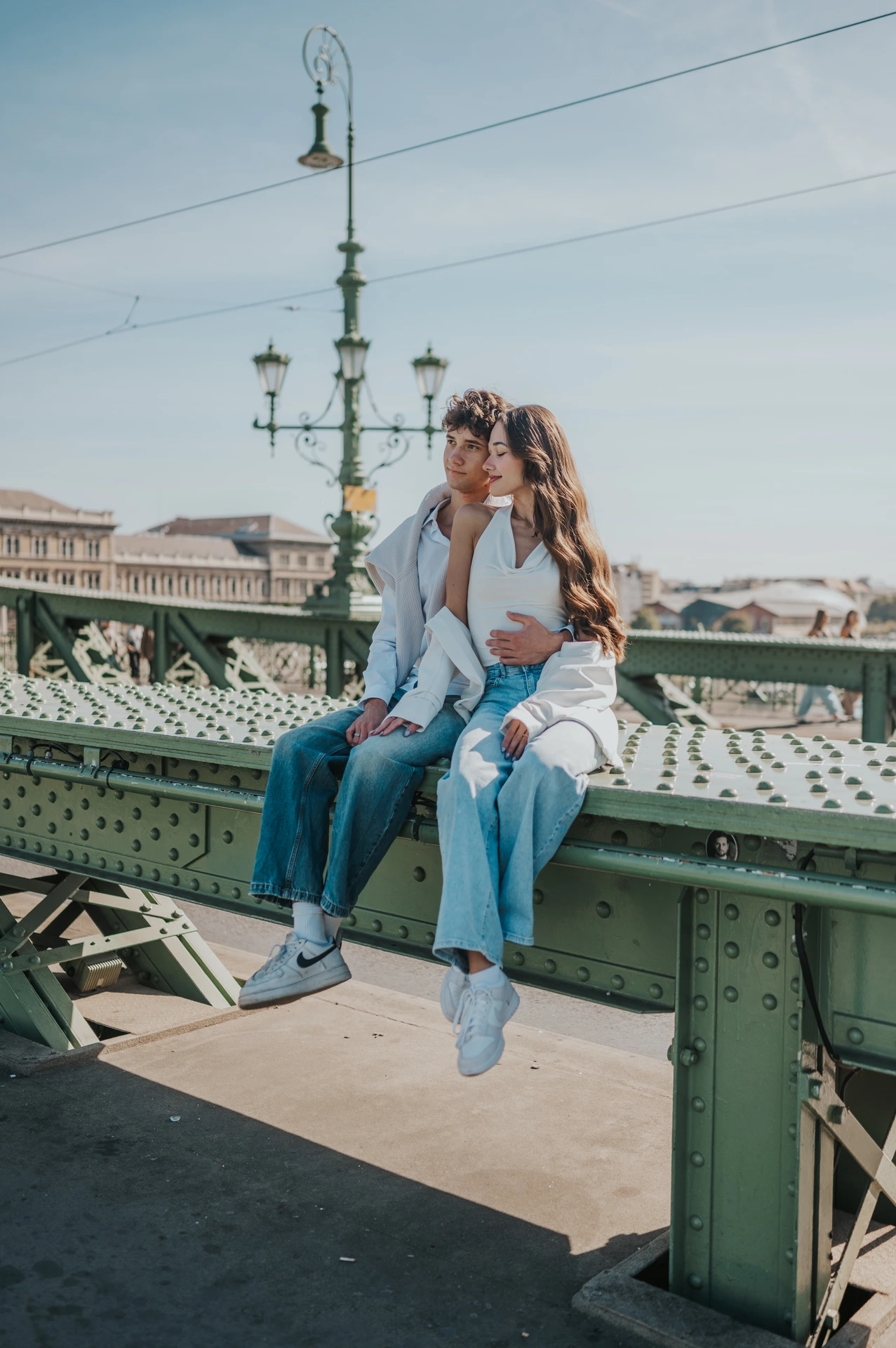 A wider portrait of the couple sitting together on the green railing during their Budapest photoshoot, with Liberty Bridge’s streetlamp and river view around them.