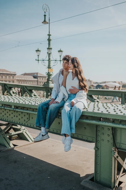 Couple photoshoot in Budapest with the Liberty Bridge railing and river view A wider portrait of the couple sitting together on the green railing during their Budapest photoshoot, with Liberty Bridge’s streetlamp and river view around them.