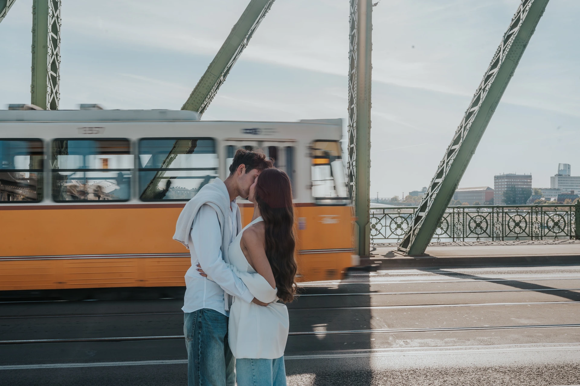 A closer view of the couple kissing during their Budapest photoshoot on Liberty Bridge, with a yellow tram moving through the background.