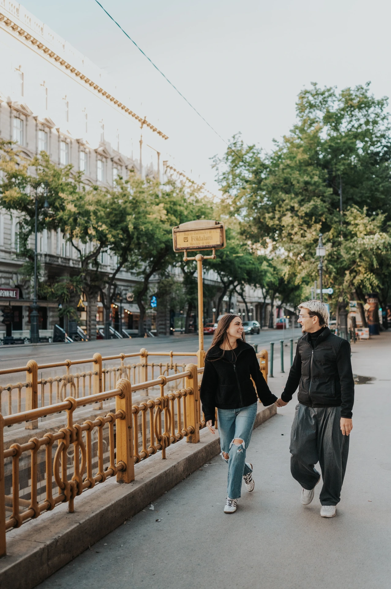 The couple walking beside the yellow railings of a metro entrance during their Budapest photoshoot, with trees and city buildings behind them.