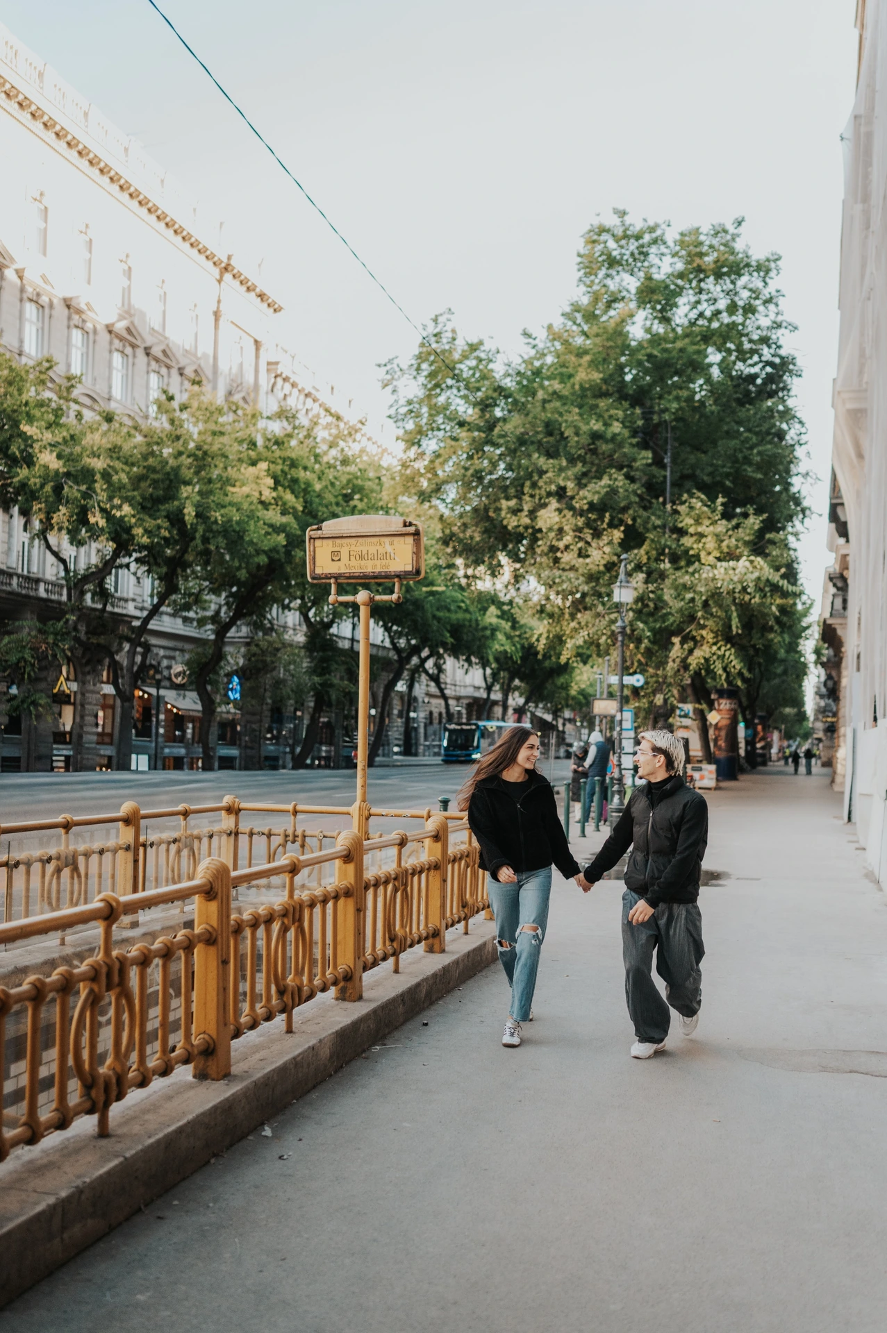 The couple walking hand in hand along the pavement beside the metro entrance during their Budapest photoshoot.