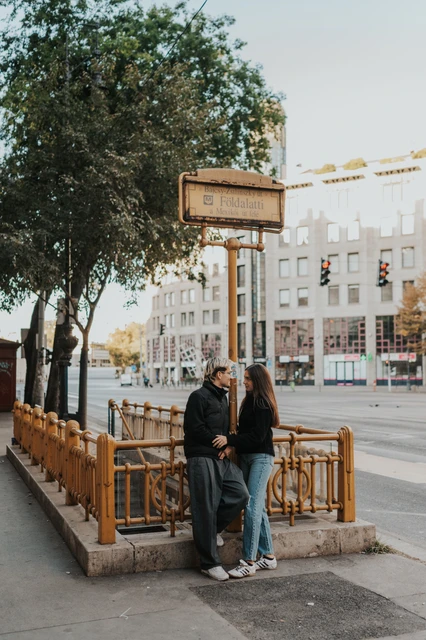 Budapest couple photoshoot at the street-level metro entrance The couple standing close together beside the yellow railings and sign at a metro entrance during their Budapest photoshoot.