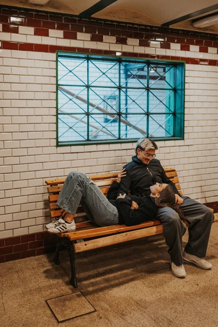 The couple relaxing together on a wooden station bench during their Budapest photoshoot, with a tiled wall and blue window behind them.