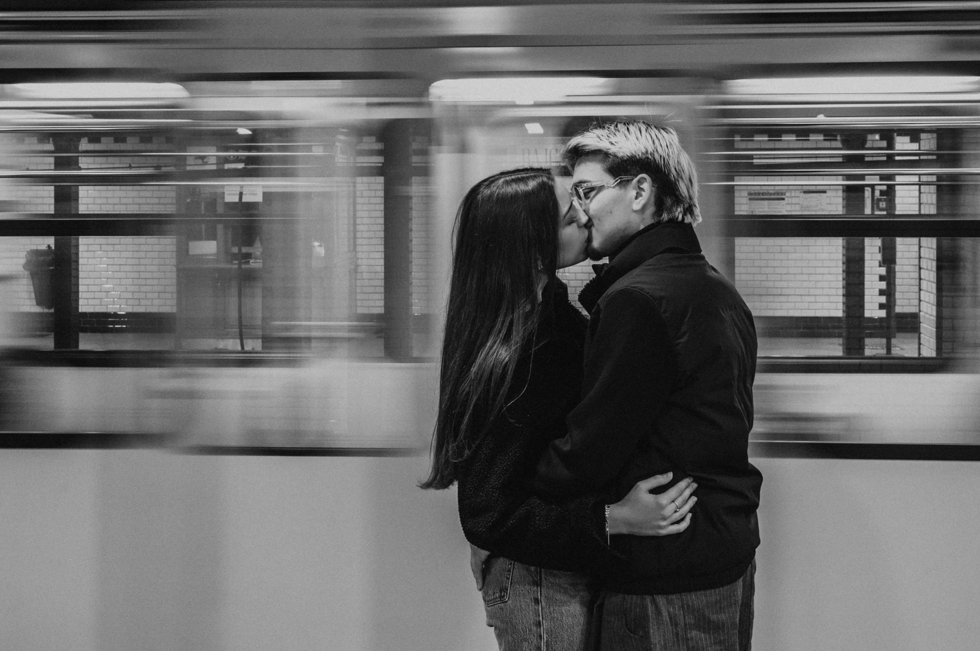 Black-and-white photograph of the couple kissing on the platform during their Budapest photoshoot, with a passing train blurred behind them.