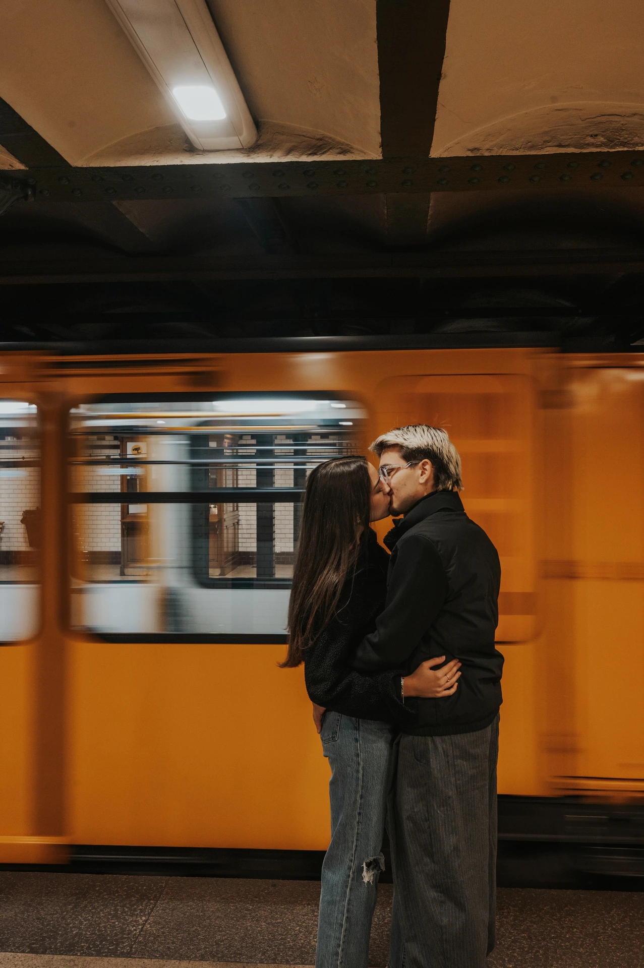 The couple holding each other close on the platform during their Budapest photoshoot, framed against a yellow metro train.