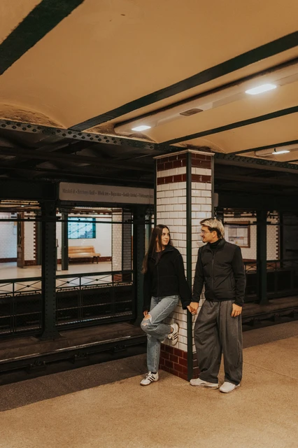 Couple photoshoot in Budapest beside the metro platform columns The couple standing beside a riveted metal column during their Budapest photoshoot in the old metro station, beneath the platform lights.