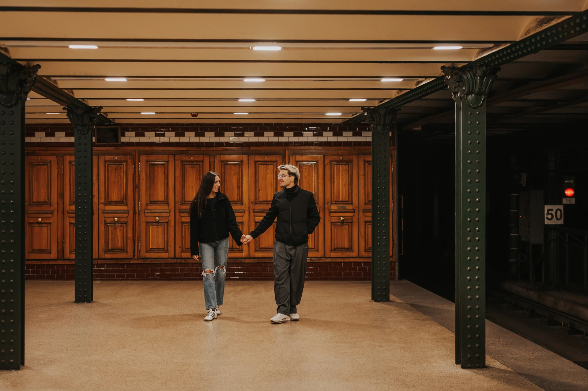 Wide view of the couple holding hands on the platform during their Budapest photoshoot, surrounded by wooden panelling and dark metal columns.