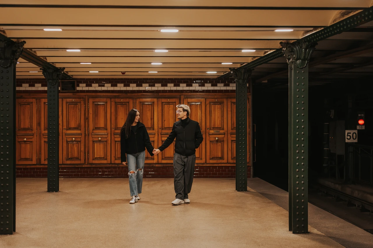 Budapest photoshoot on a platform with wooden panelling Wide view of the couple holding hands on the platform during their Budapest photoshoot, surrounded by wooden panelling and dark metal columns.