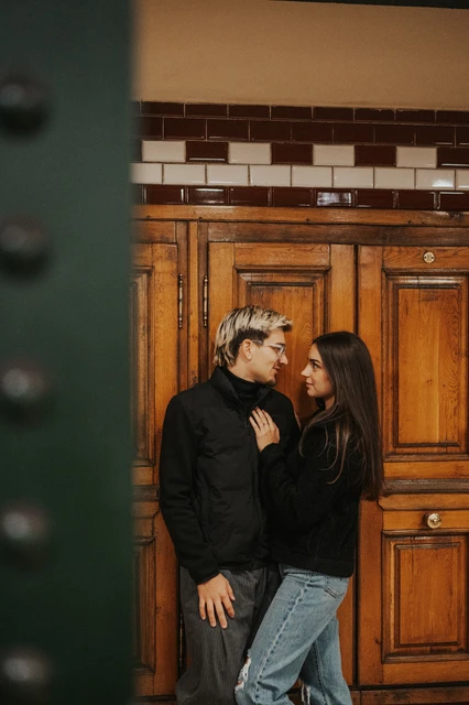 Photoshoot in Budapest beside the wooden metro station doors The couple leaning close beside wooden station doors during their Budapest photoshoot, with a dark metal column in the foreground.