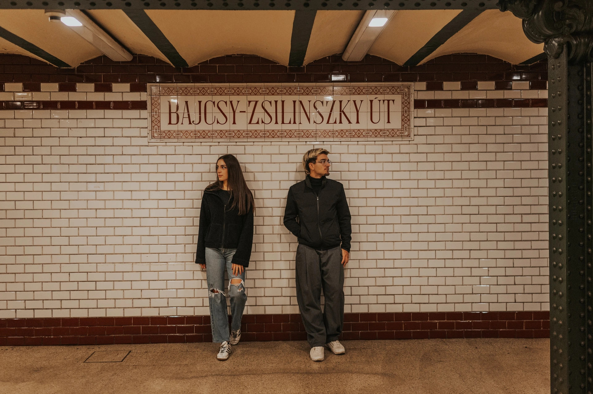 Wide portrait of the couple standing beneath the station name sign during their Budapest photoshoot, against a wall of cream tiles.