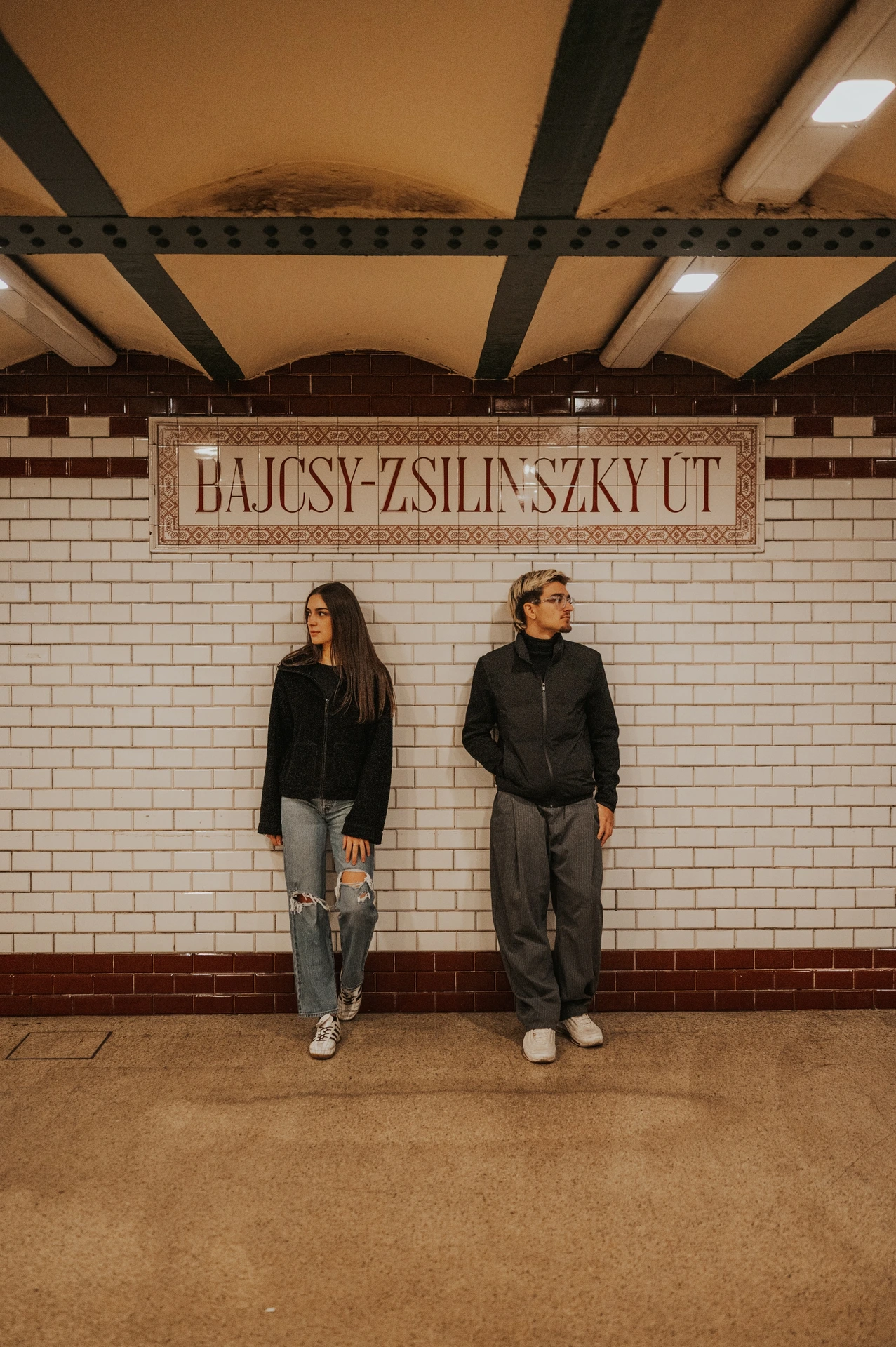 The couple standing side by side against the tiled station wall during their Budapest photoshoot, looking in different directions beneath the sign.