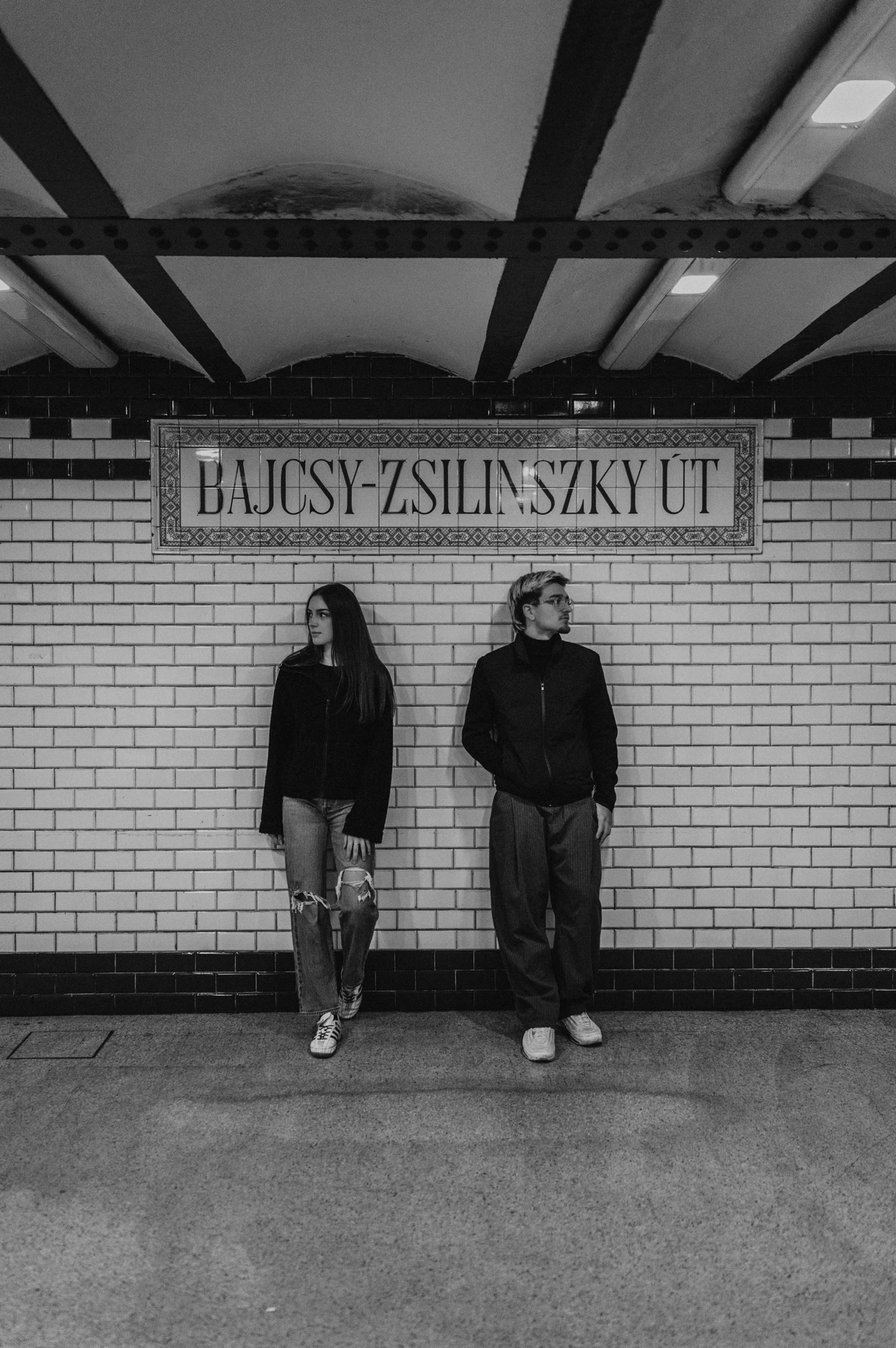 Black-and-white portrait of the couple standing beneath the station sign during their Budapest photoshoot, with tiled walls and ceiling beams above them.