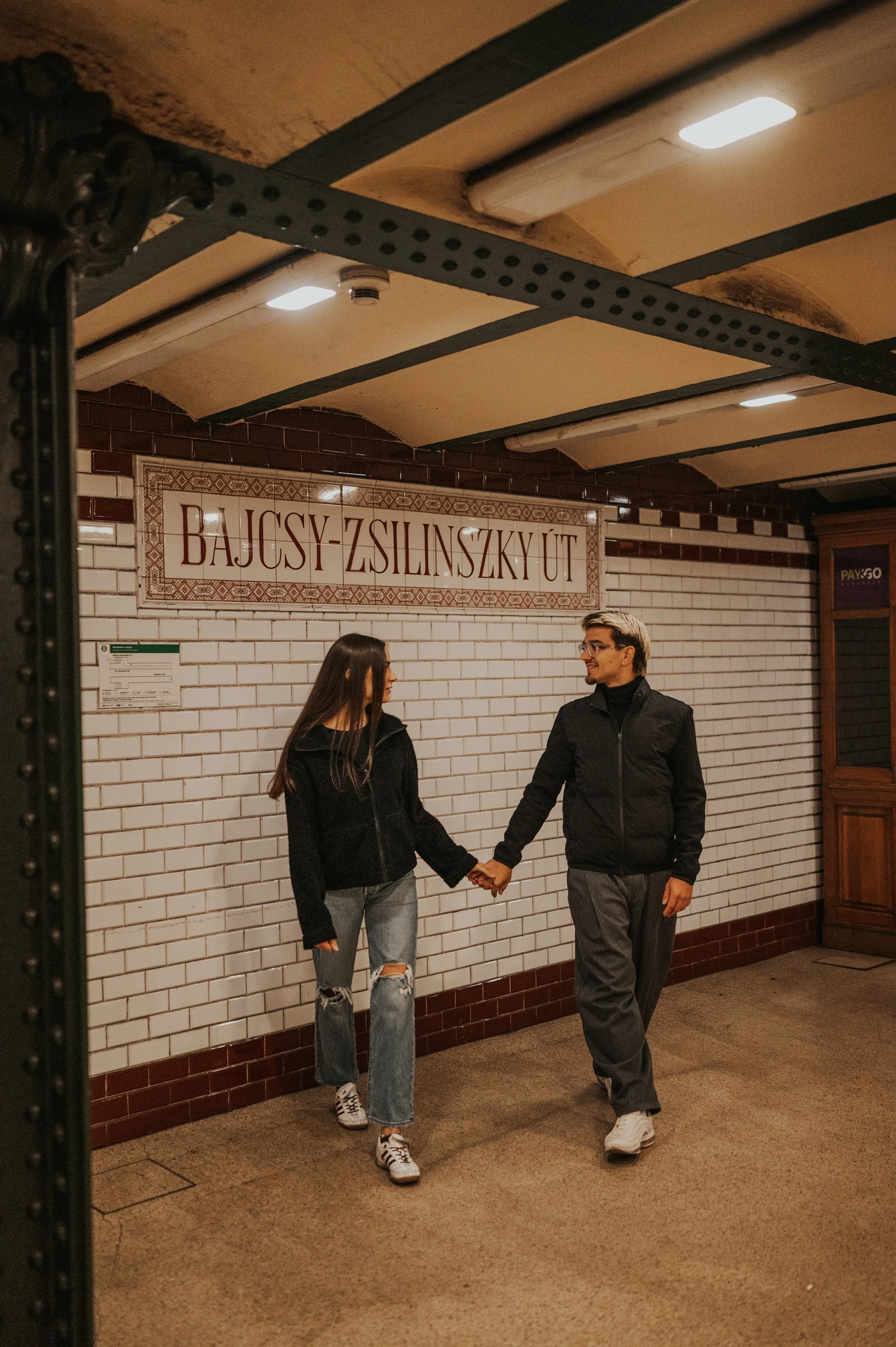 The couple walking hand in hand along the tiled station wall during their Budapest photoshoot in the old metro.