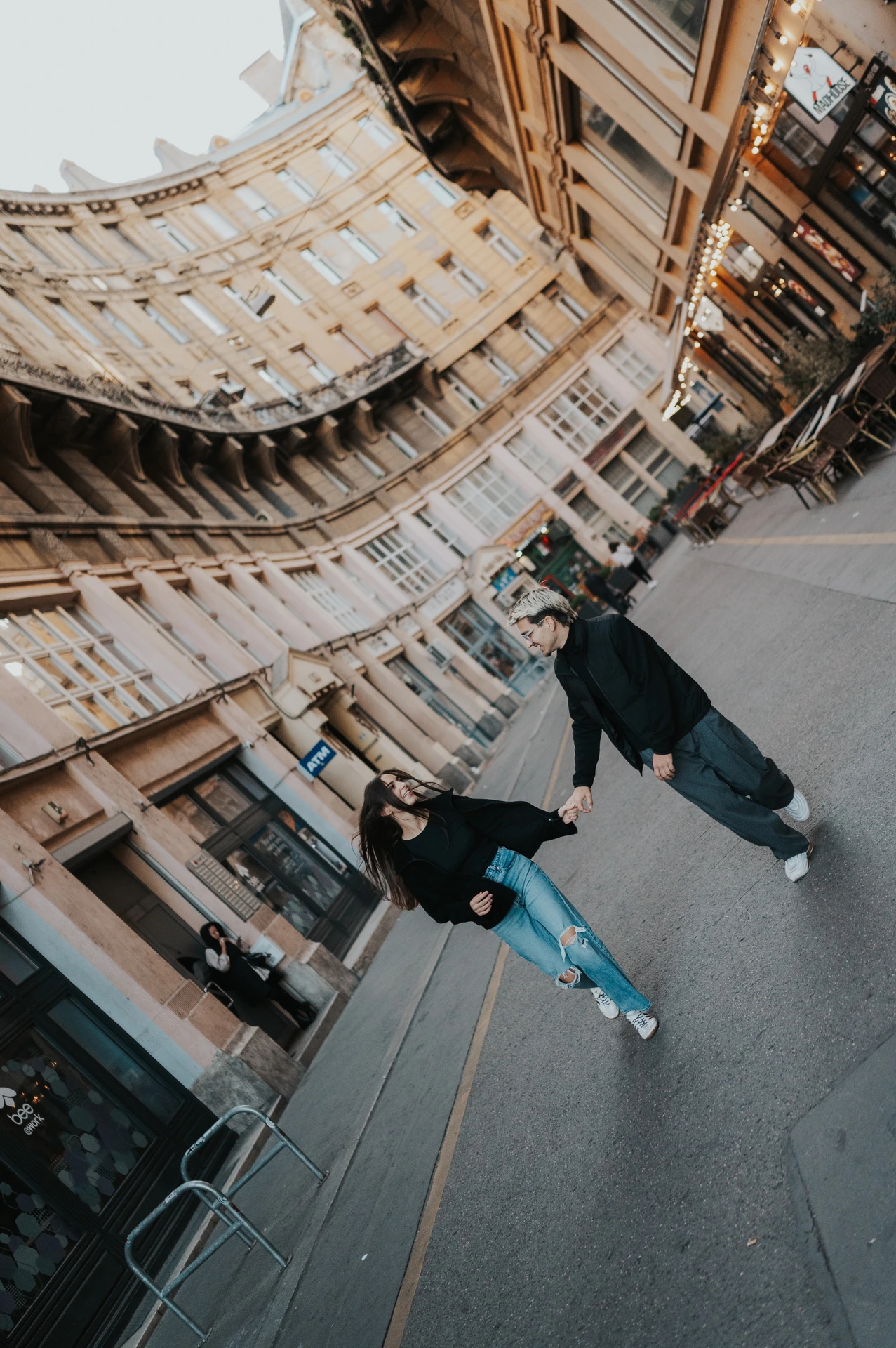 A tilted street photograph of the couple moving playfully hand in hand during their Budapest photoshoot, with a curved city building behind them.