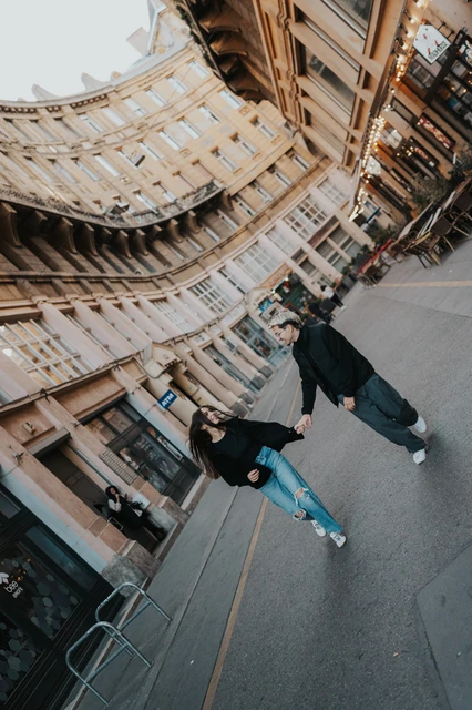Playful Budapest couple photoshoot on the city streets A tilted street photograph of the couple moving playfully hand in hand during their Budapest photoshoot, with a curved city building behind them.