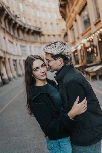 Budapest couple photoshoot sharing a kiss on the street The man kissing his partner beside the street during their Budapest photoshoot, with her looking towards the camera and a curved building behind them.