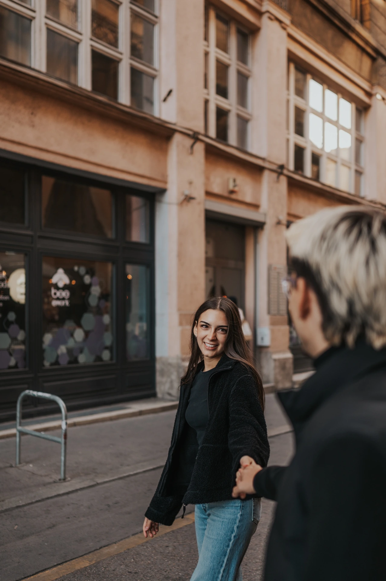 The woman glancing back towards her partner during their Budapest photoshoot on a city street, with him softly out of focus in the foreground.