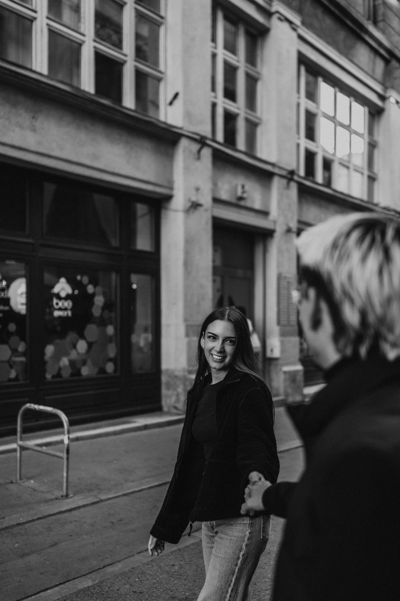 Black-and-white photograph of the woman looking back towards her partner during their Budapest photoshoot, with city façades behind her.