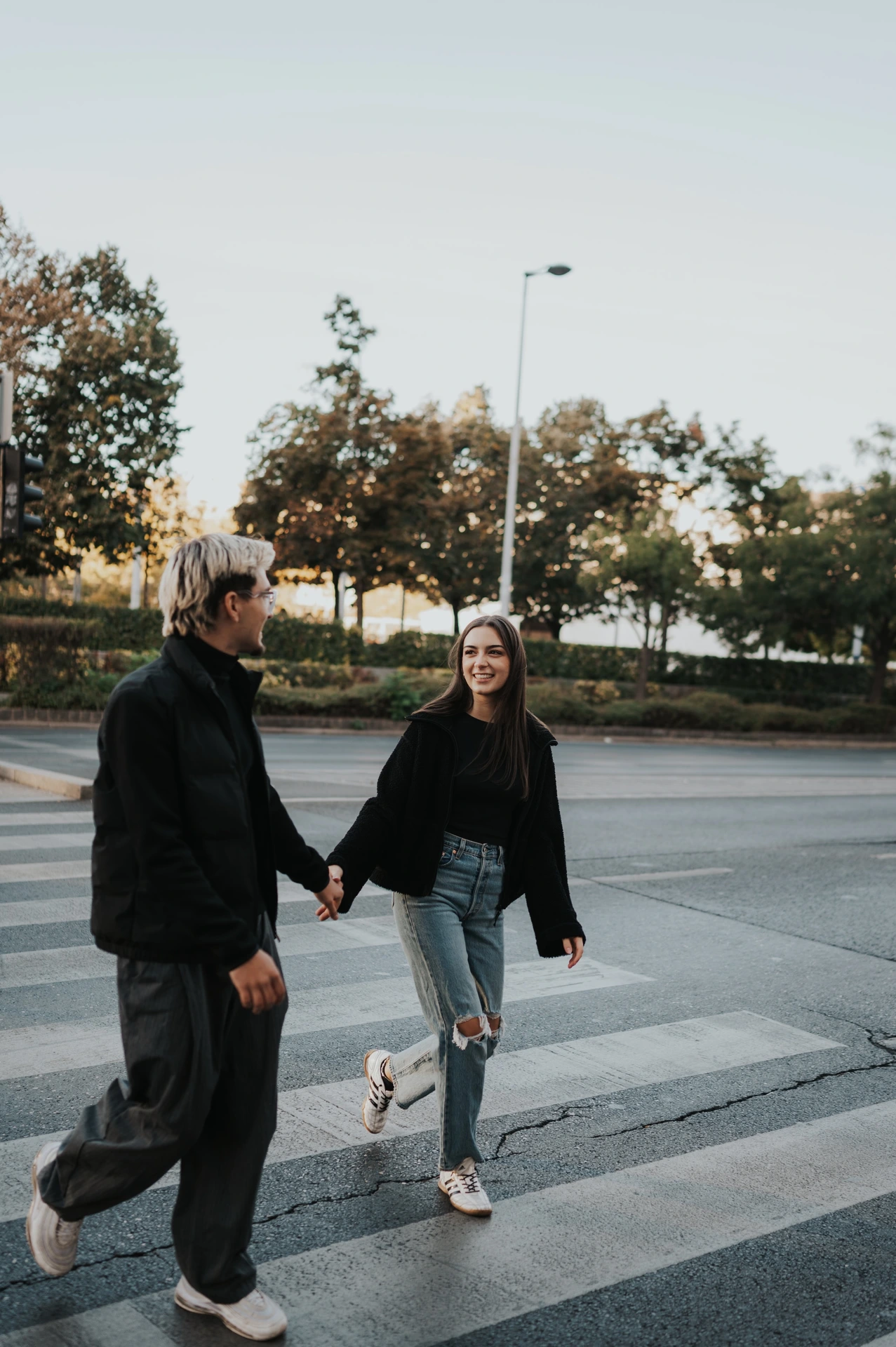 The couple crossing a city street together during their Budapest photoshoot, wearing dark jackets and casual trousers.