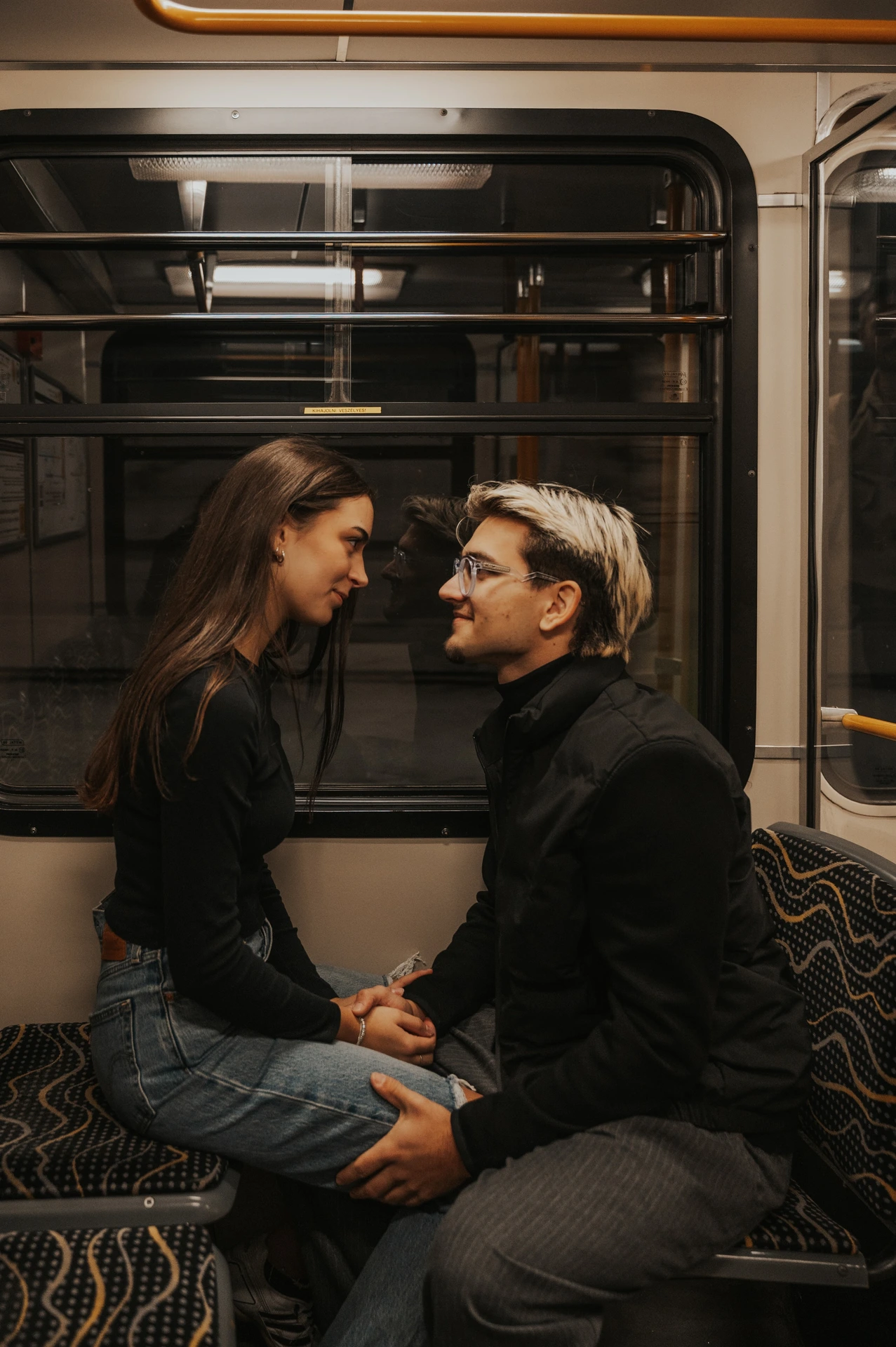 The couple seated close together inside the metro during their Budapest photoshoot, holding hands beside the window and patterned seats.