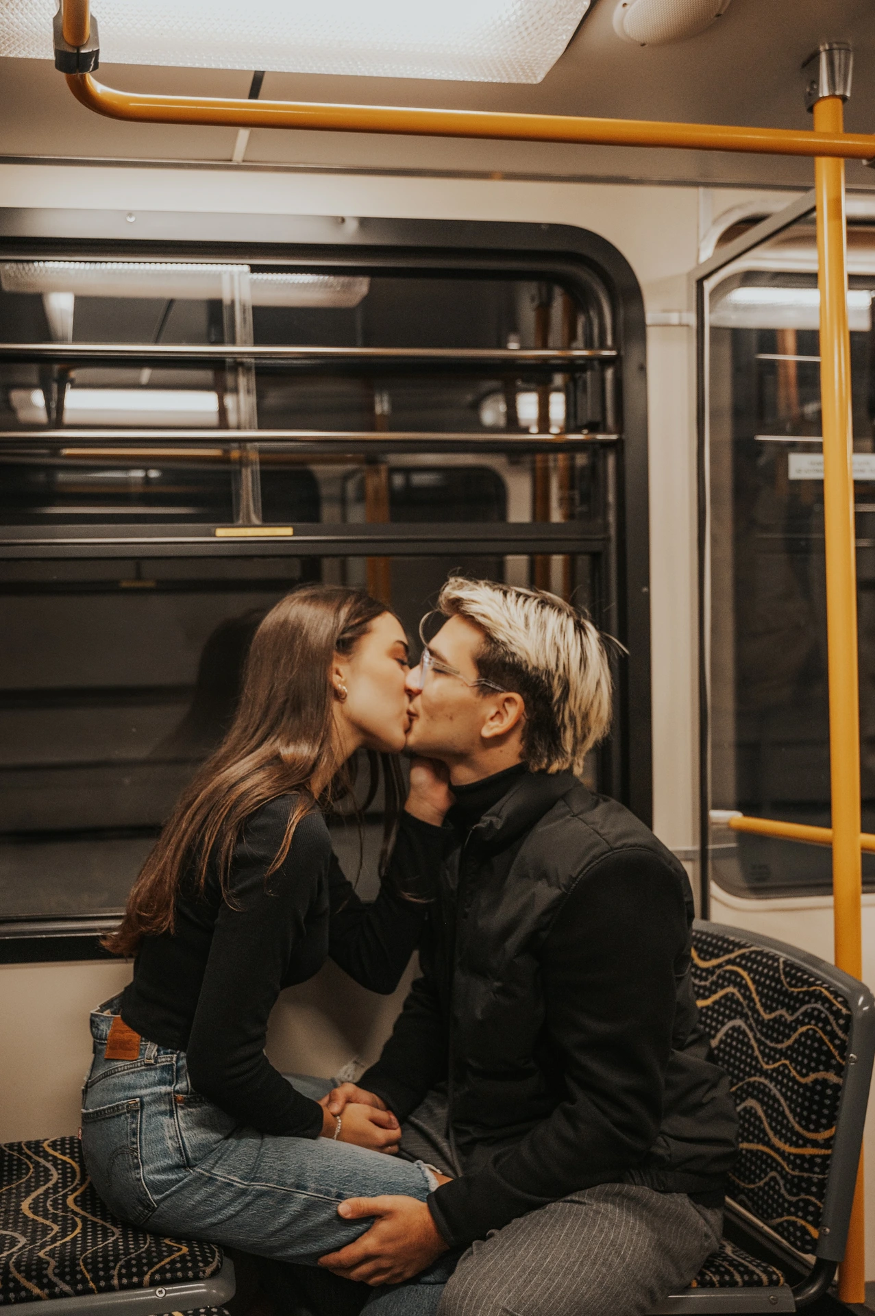 The couple kissing while seated beside a metro window during their Budapest photoshoot, with a yellow handrail and patterned seats around them.