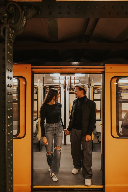 The couple standing hand in hand inside a yellow metro carriage during their Budapest photoshoot, photographed through the open doors.