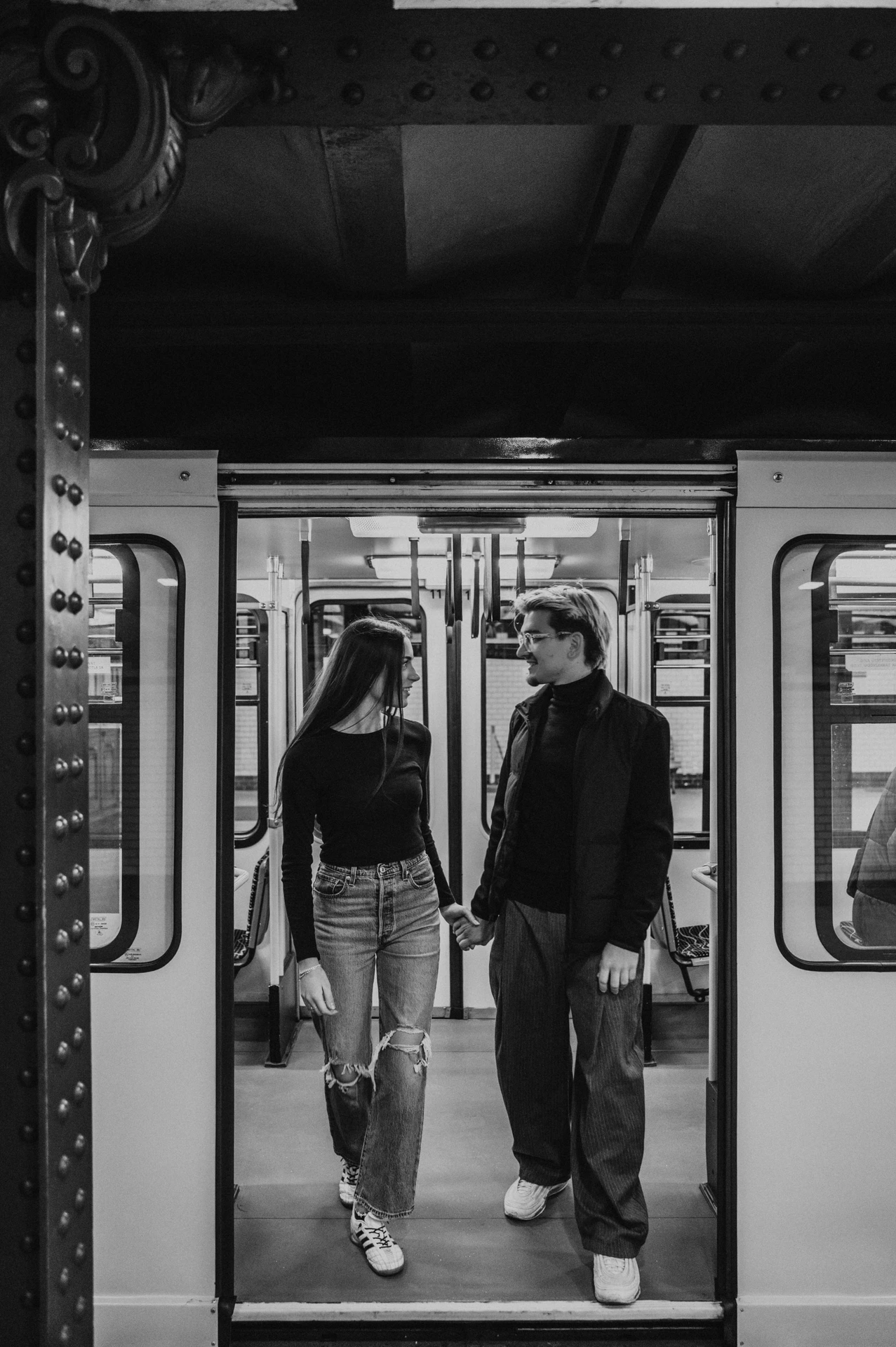 Black-and-white photograph of the couple holding hands inside the metro during their Budapest photoshoot, viewed through the open carriage doors.