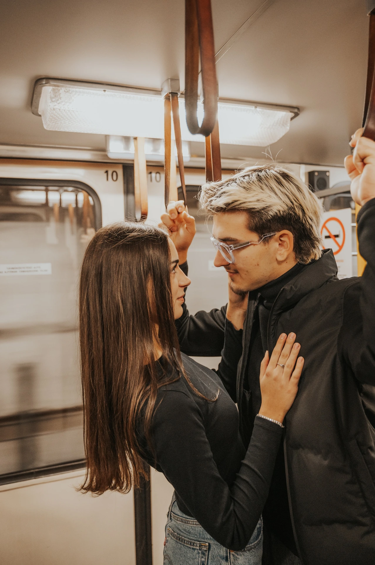 The couple facing each other beside the metro window during their Budapest photoshoot, with him holding an overhead rail.