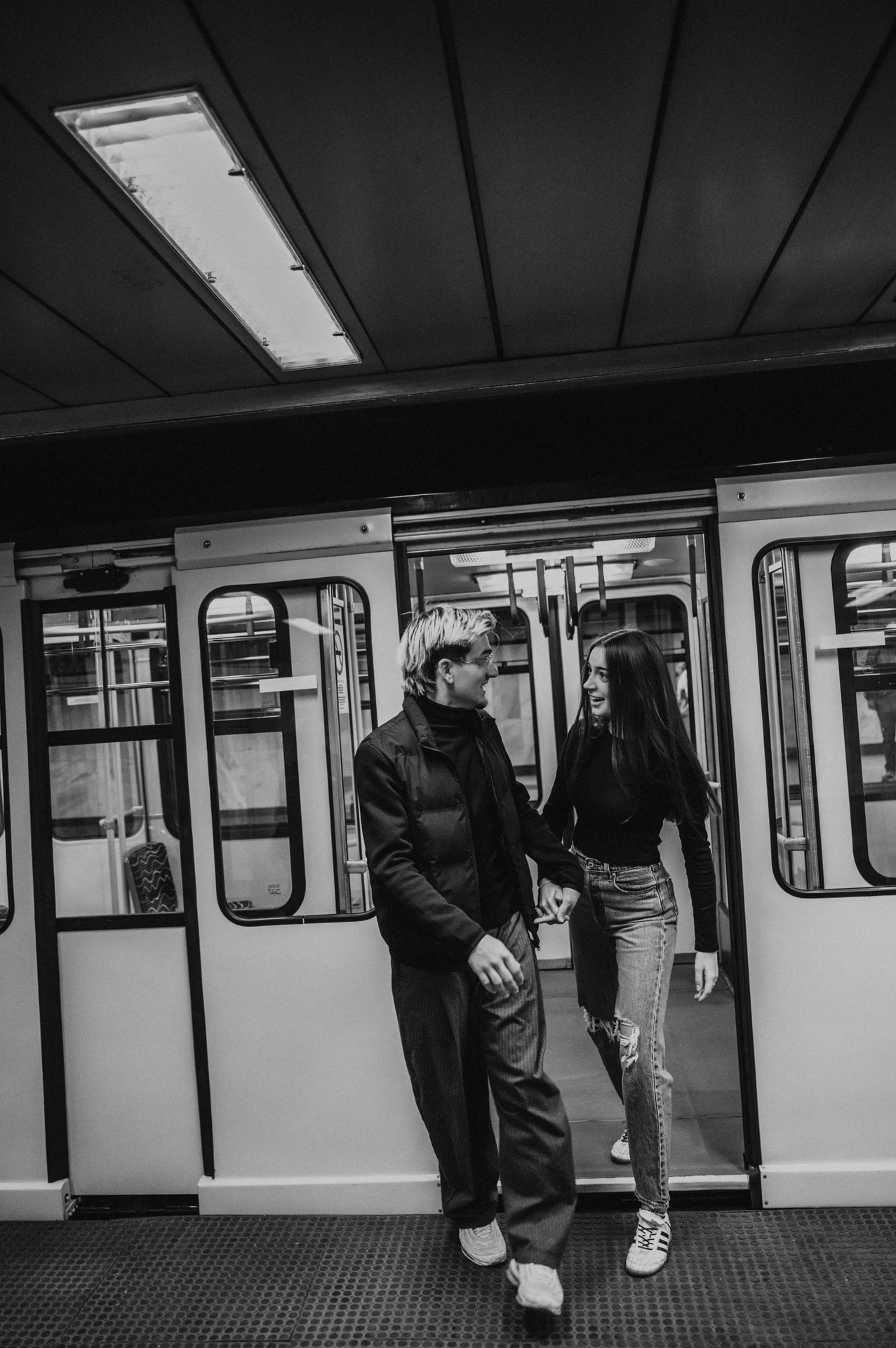 Wide black-and-white view of the couple standing close together inside the metro during their Budapest photoshoot, surrounded by carriage doors and ceiling lights.