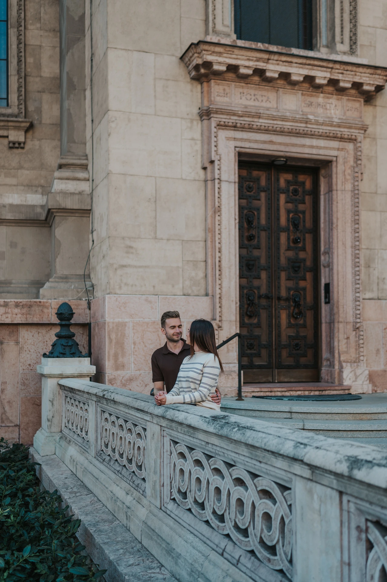 The couple leaning close beside a carved stone balustrade and doorway at St Stephen’s Basilica during a couple photoshoot in Budapest.