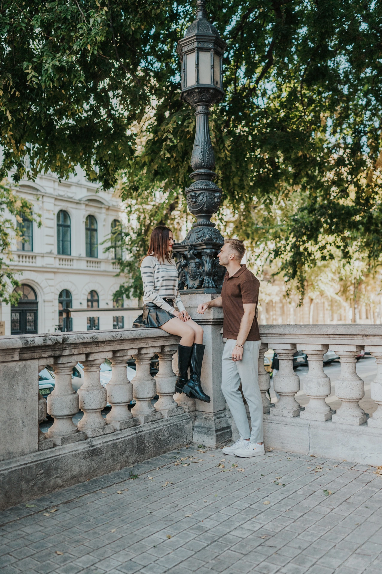 The couple beside a carved stone balustrade and green lamp beneath trees near St Stephen’s Basilica during a couple photoshoot in Budapest.