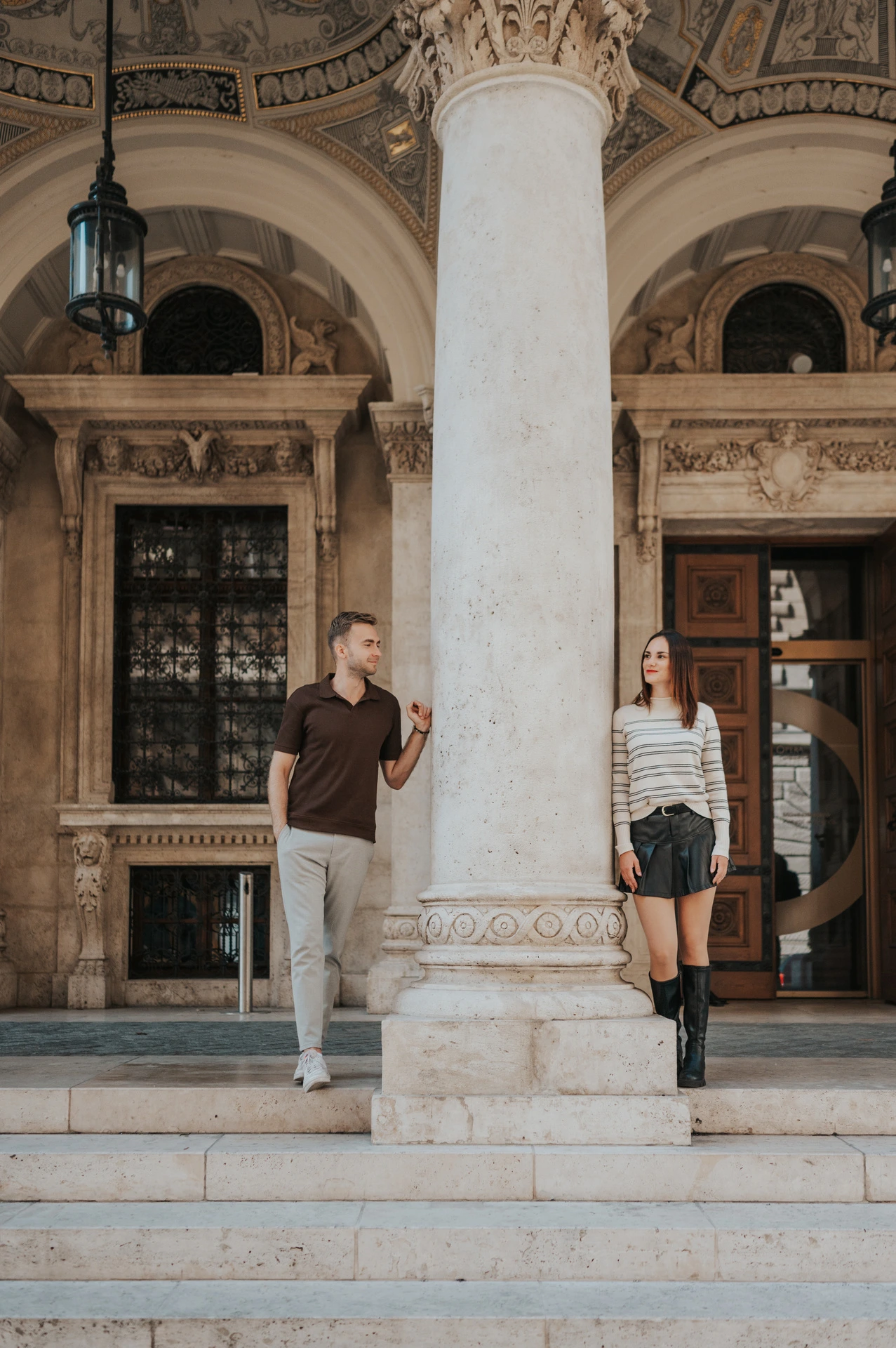 The couple standing on opposite sides of a tall stone column above the Opera steps during a couple photoshoot in Budapest.