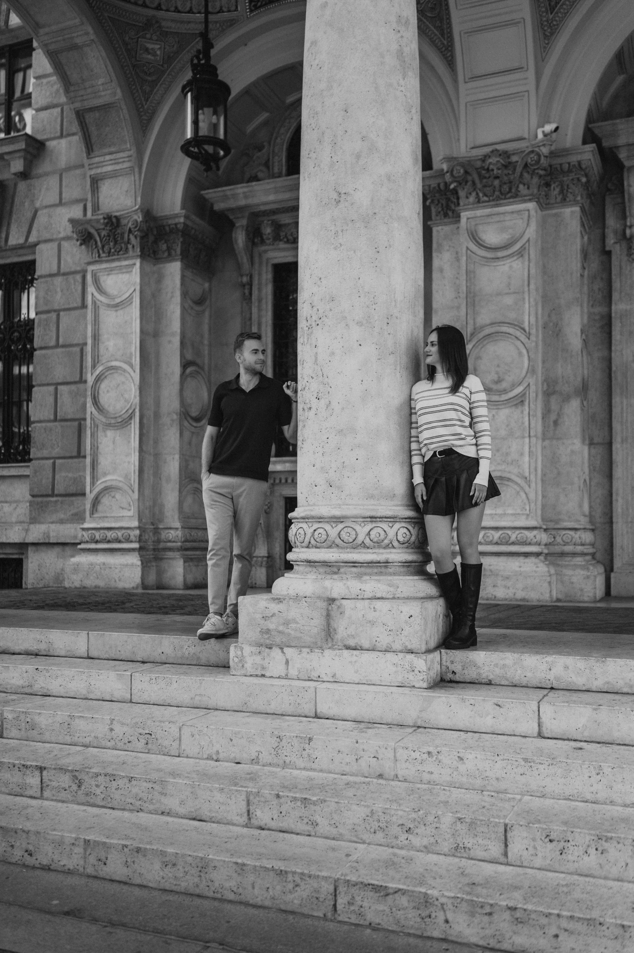 Black-and-white photograph of the couple standing beside a large stone column at the Opera entrance during a couple photoshoot in Budapest.