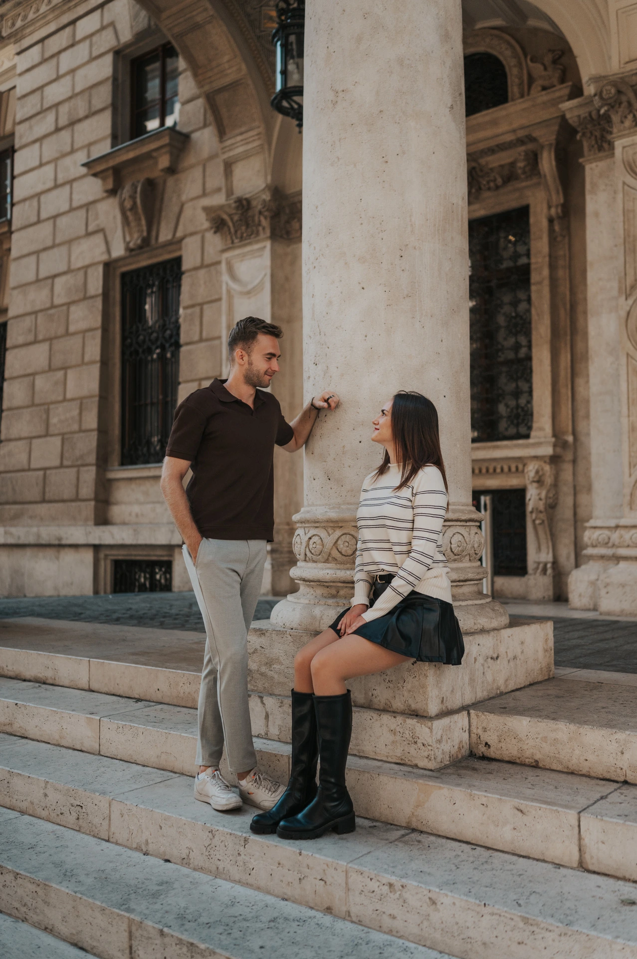 The woman sitting on the Opera steps while her partner leans beside a stone column during a couple photoshoot in Budapest.