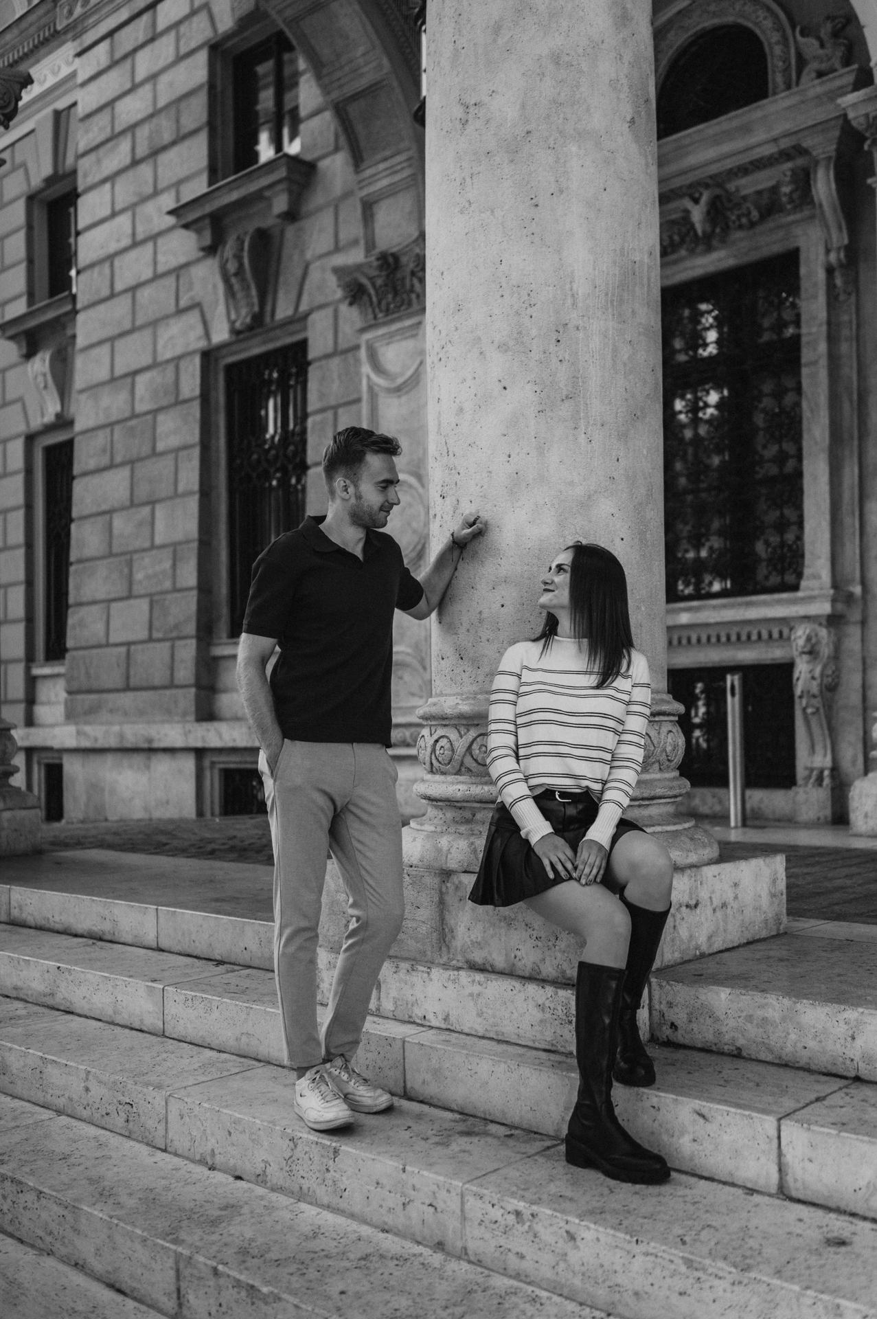 Black-and-white photograph of the woman sitting on the Opera steps beside her partner and a large stone column during a couple photoshoot in Budapest.