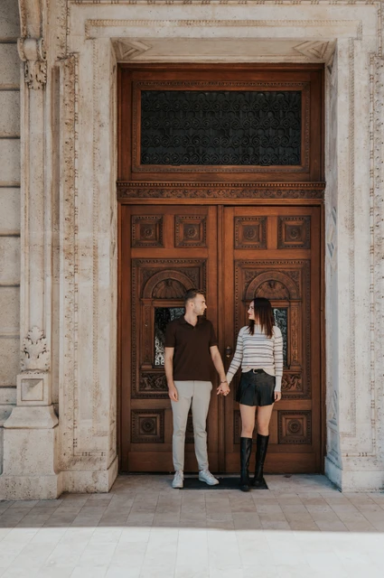 The couple holding hands in front of the Opera’s wooden doors and pale stone surround during a couple photoshoot in Budapest.