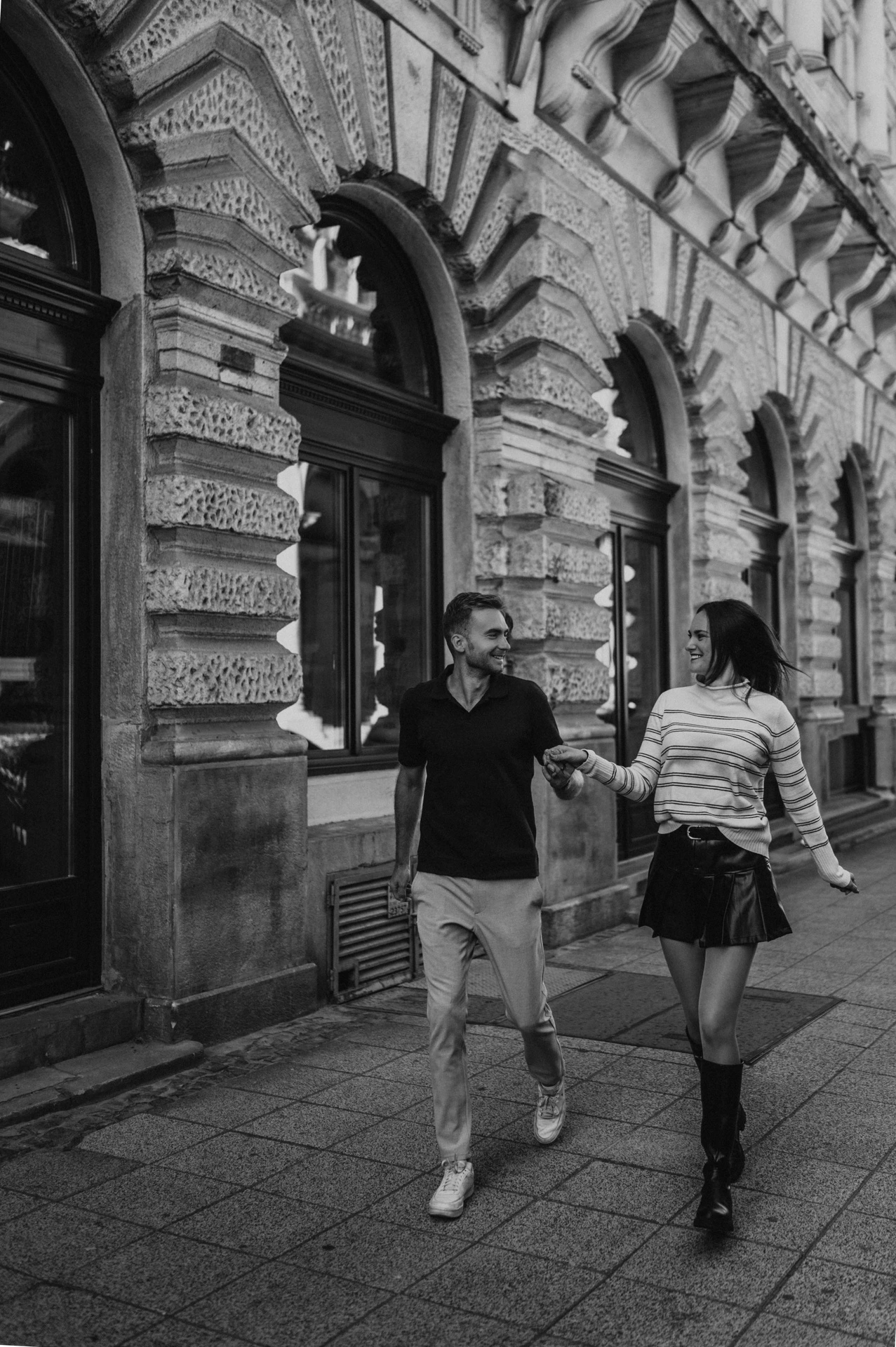 Black-and-white photograph of the couple walking along the pavement beside the Opera’s carved façade during a couple photoshoot in Budapest.