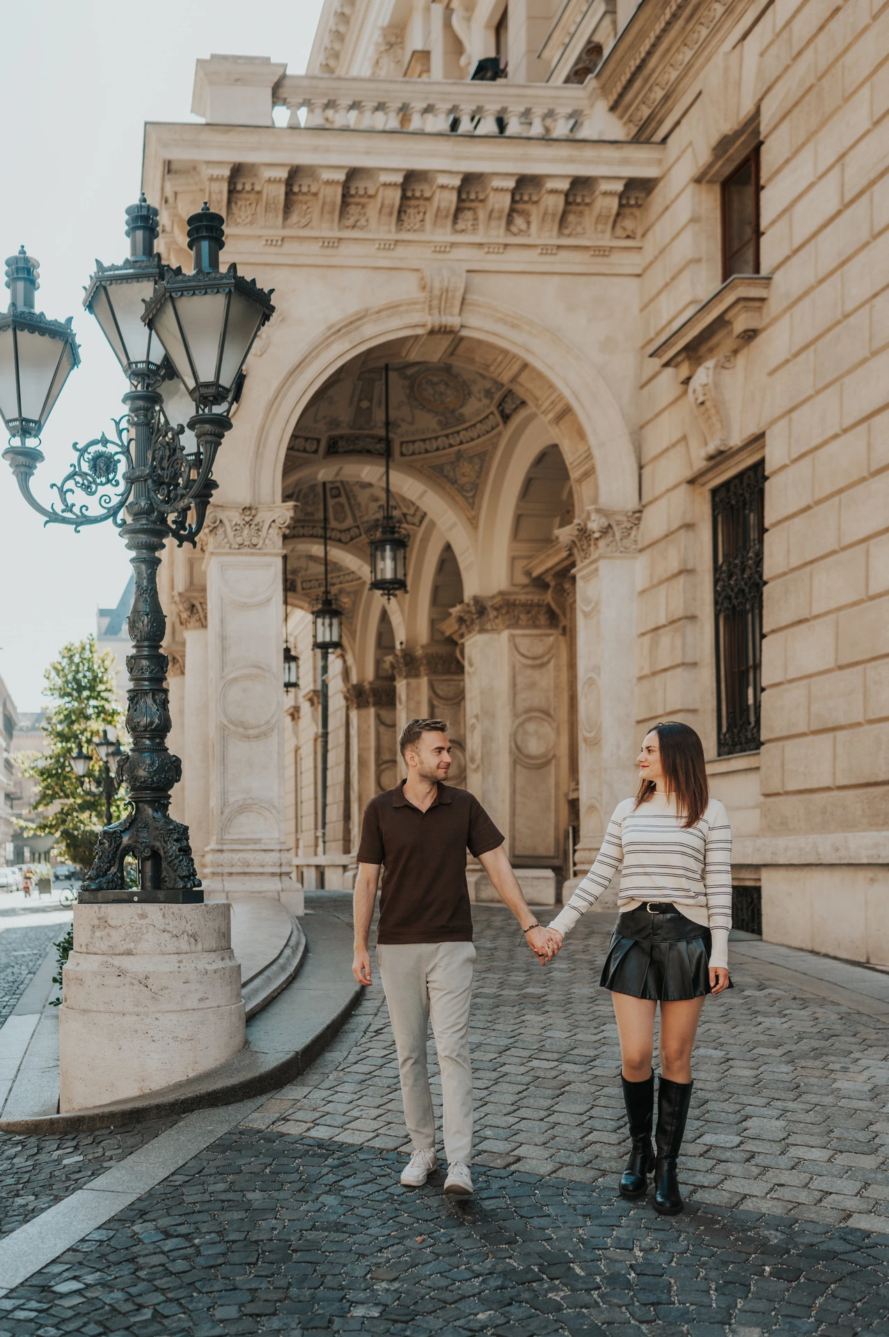The couple holding hands beside the Opera’s arched entrance and ornate green lamp during a couple photoshoot in Budapest.