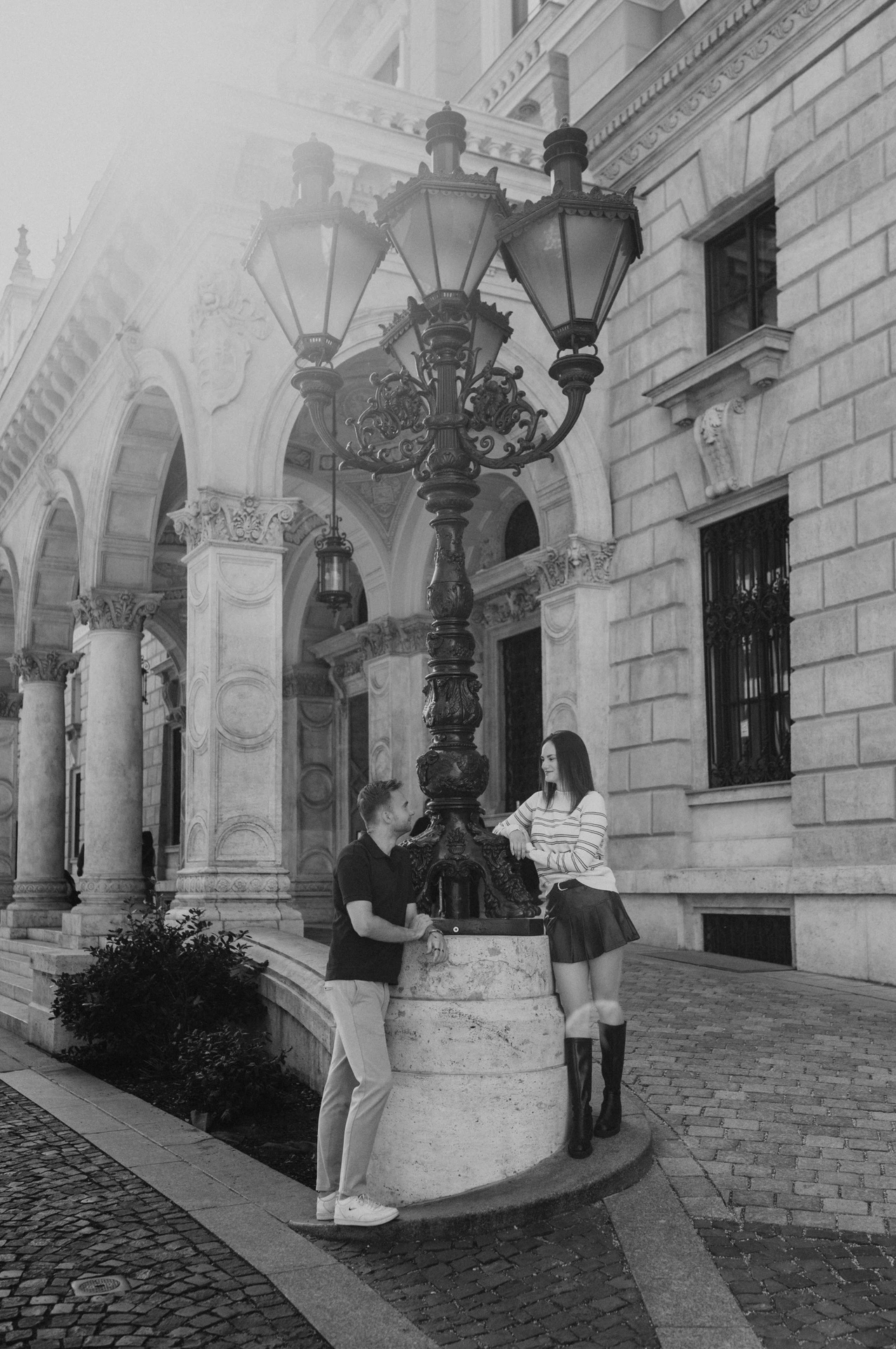 Black-and-white photograph of the couple standing on opposite sides of an ornate street lamp outside the Opera during a couple photoshoot in Budapest.