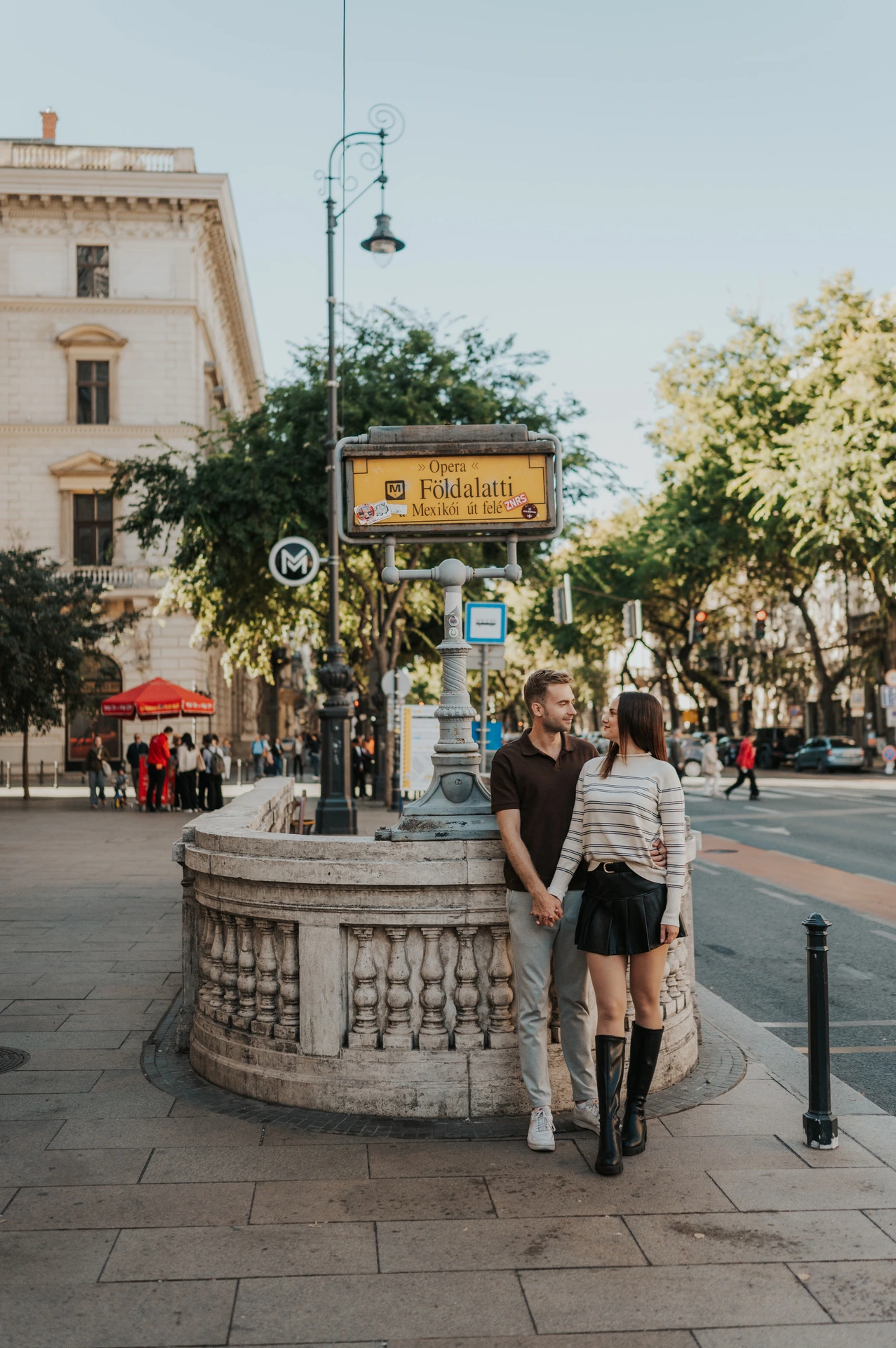 The couple standing together beside the stone balustrade and yellow sign at Opera metro station during a couple photoshoot in Budapest.