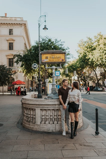 The couple standing together beside the stone balustrade and yellow sign at Opera metro station during a couple photoshoot in Budapest.